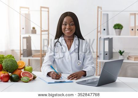 Smiling Black Woman Dietologist Working With Laptop In Modern Office, Having Fresh Fruits And Vegeta