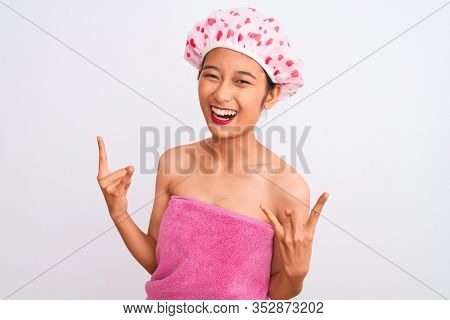 Young chinese woman wearing shower towel and cap bath over isolated white background shouting with crazy expression doing rock symbol with hands up. Music star. Heavy concept.