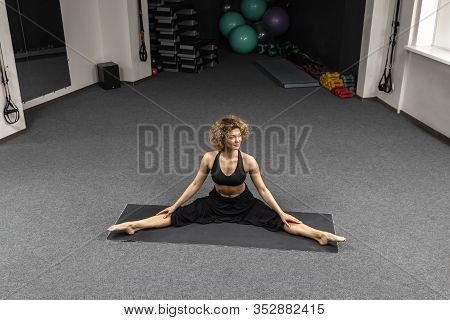Sporty Woman In Black Clothing Practicing Yoga, Doing Splits Stretching Legs On Mat At Gym. Sport Mo