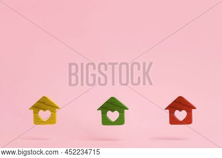 Three Little Multi-colored Wooden Houses With A Carved Heart Inside. Pink Background With Copy Space