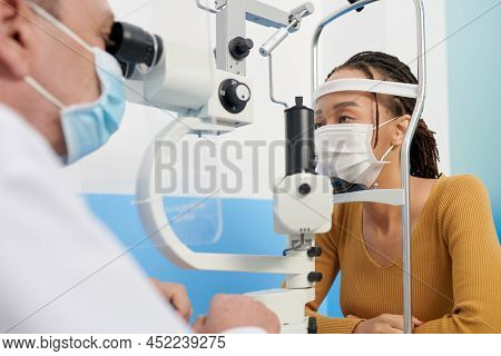 Black Woman In Medical Mask Getting Eyes Examined With Autorefractometer To Get Lens Prescription