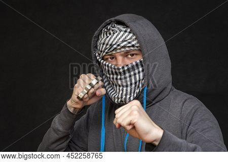 Brass knuckles in the hand of a man with a scarf covering his face on a dark background, selective f