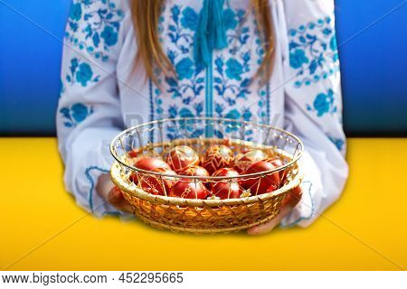 Defocus Ukrainian Girl In Vyshyvanka Holding Basket Of Colored Red Eggs On Blue And Yellow Backgroun
