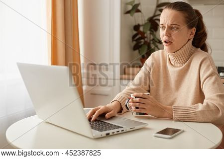 Shocked Estonished Caucasian Woman Wearing Beige Jumper Posing In Kitchen And Working On Laptop, Loo