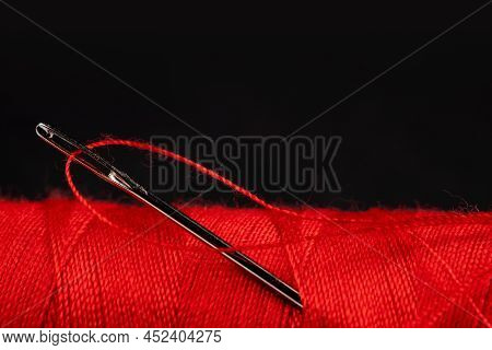Eye Of The Needle With Red Polyester Thread. Spool Of Red Polyester Sewing Thread In The Background.