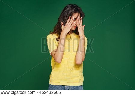 Scared Or Embarrassed Young Girl Covering Eyes By Hands, Peeping On Green Studio Background With Cop