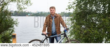 An Adult Blond Man Brown Corduroy Jacket Is Standing By The River In The Park With A Bicycle.