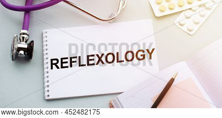 The Word Reflexology Written In Notepad With Sthethoscope, Pills And Pen Lying On Doctor's Table.