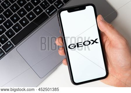 Geox App Logo On The Iphone Screen For Online Shopping With A Laptop On The Background. November 202