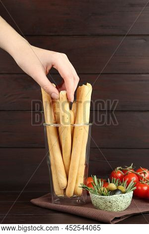 Woman Taking Fresh Delicious Grissino From Glass At Wooden Table, Closeup
