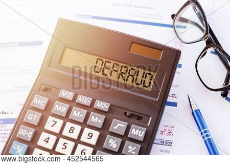 Defraud Is A Word Written On Calculator, Lying On White Surface Next To Glasses And A Blue Pen.