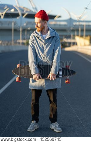 Casual Man With Longboard On Sunset Over Modern City Building. Hipster Skater Holding Skateboard Wea
