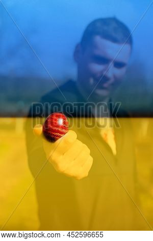 Defocus Ukrainian Young Man Holding One Colored Red Egg On Nature Background. Easter, Ukraine Flag. 