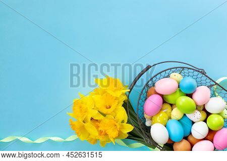 Basket With Easter Eggs And Bouquet Of Yellow Narcissus On Blue Paper Background. Flat Lay  Top View