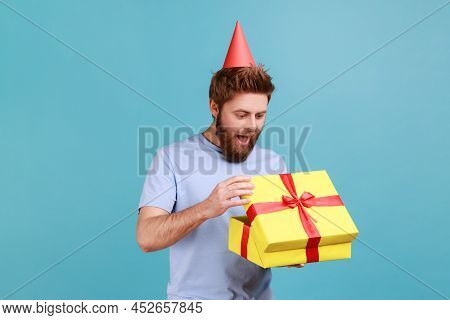 Man With Party Cone Hat Holding Opened Gift Box And Looking At Present With Excited Look, Unpacking 