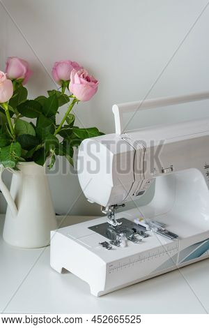 White Modern Ewing Machine And Its Components Near Pink Roses In Vase. Sewing Needle Plate And Repla