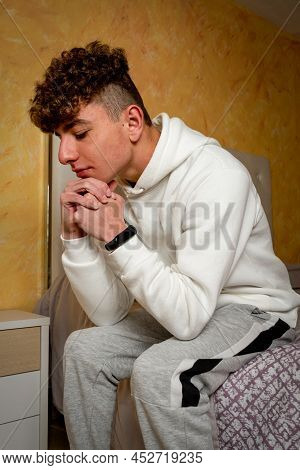 Caucasian Young Man With Curly Hair Sitting On His Bed With A Worried Gesture, He Analyzes The Probl