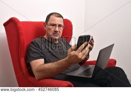 Middle-aged Man With A Laptop On His Lap Sed In A Red Armchair. The Man Is Holding Two Smartphones