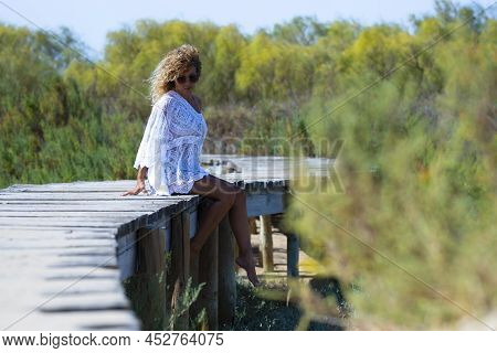 Attractive Woman In White Dress Sitting Barefoot On Elevated Footbridge Leading Towards Natural Fore