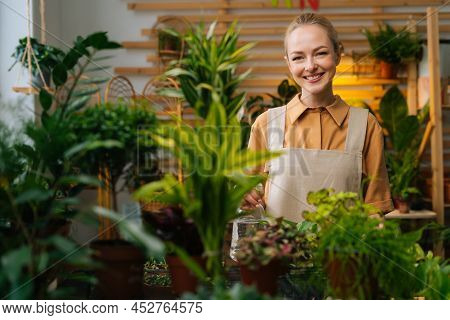 Portrait Of Cheerful Young Woman In Apron Spraying Water On Houseplants In Flower Pots By Sprayer, L