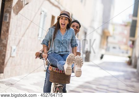 Joyful Couple Riding Bicycle Outdoors In City. Man With Delighted Woman Sitting On Handlebar While R