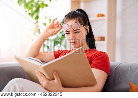 A Young Brunette With Glasses Is Reading A Book, Having Poor Eyesight At Home. A Student Experiencin
