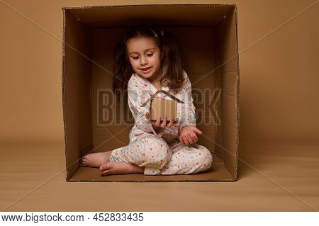 Adorable Caucasian Little Girl Hiding Inside A Box And Holding A Craft Cardboard House Model. Real E