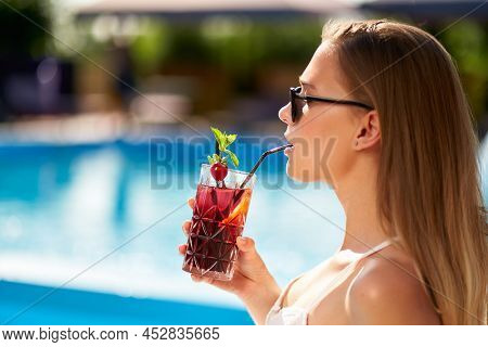 Blonde Woman Drinks Refreshing Cocktail Sunbathing And Sitting Near Swimming Pool At Tropical Spa. F