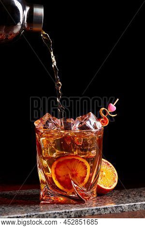 Alcohol Cocktail Pouring From Shaker Into Gless With Orange Peel Ice On Stone Desk.