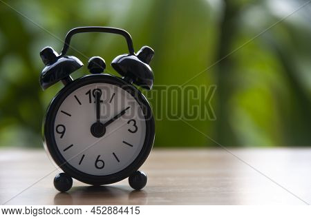 Black Alarm Clock Isolated On Blurred Nature Background. The Clock Set At 2 O'clock. Copy Space.