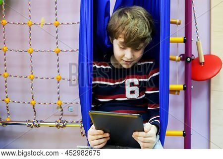 Child Boy Watching Cartoons On A Tablet While Sitting In A Hammock At A Children's Center For The De