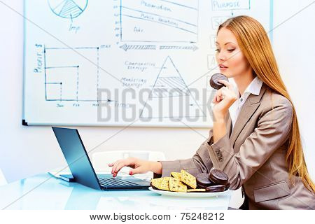 Beautiful businesswoman working in the office with a laptopn and eating cookies.