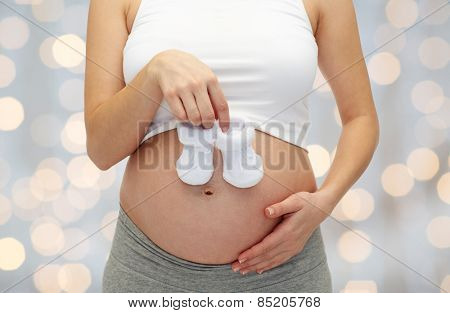 pregnancy, motherhood, people and expectation concept - close up of happy pregnant woman touching her bare tummy and holding babys bootees over holiday lights background
