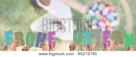 Hands holding up frohe ostern against little girl sitting on grass showing basket of easter eggs to camera