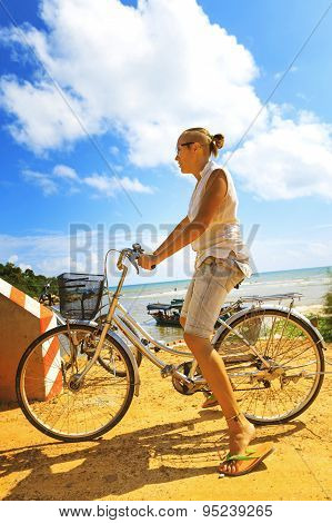 Young Woman Riding Bicycle Across River Bridge Next To Tropical Park