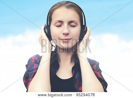 Portrait Of Young Woman In Headphones Relaxes And Listens To Music Over Blue Sky With Clouds