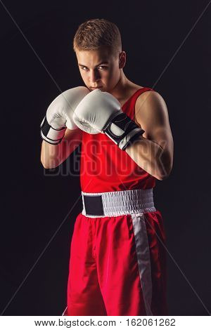 Young handsome boxer sportsman in red boxer suit and white gloves standing on black backgound. Copy space.
