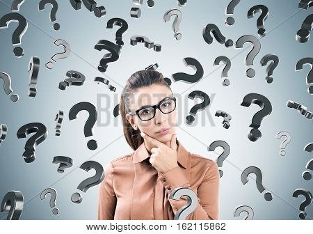 Thinking woman in glasses with her fingers on the chin is standing near a gray wall with question marks.