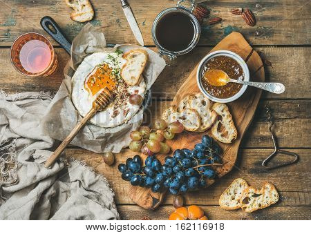 Cheese, fruit and wine set. Camembert in small pan, honey, fig jam in jar, persimmon, grapes, pecan nuts, grilled baguette slices and glass of rose wine over rustic wooden background, top view