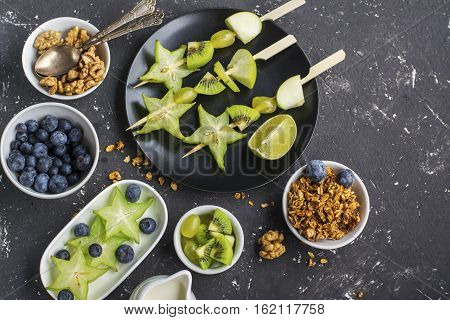Healthy fresh breakfast. Granola, quinoa bowl, various nuts and fruit skewers with green fruits on a dark background. Top view. Color year. Greenery. Top