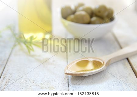 Spoon with olive oil next to an oilcan and some natural green olives. Scene on a wooden white table of a rustic kitchen.