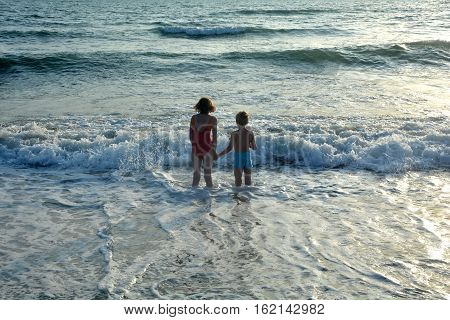 Boy and girl stand holding hands in the water on the beach and look at the sea at the sundown