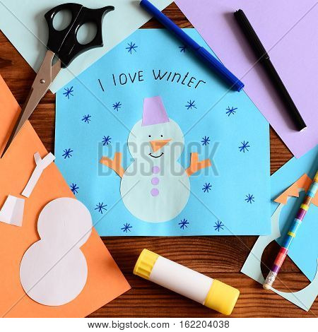 Child made a paper card with a snowman, snowflakes and words I love winter. Scissors, glue stick, pencil, markers, colored paper sheets on a wooden table. Children paper crafts. Closeup. Top view