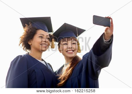 education, graduation, technology and people concept - group of happy international students in mortar boards and bachelor gowns taking selfie by smartphone outdoors