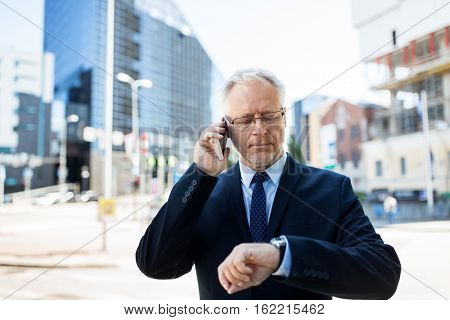 business, technology, time, punctuality and people concept - senior businessman calling on smartphone an looking at wristwatch or smart watch on his hand in city