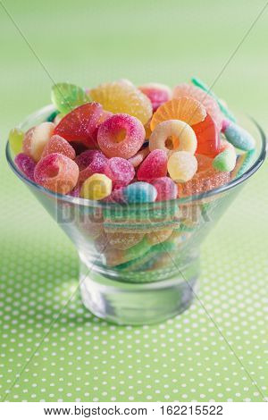 Glass bowl filled with brightly colored sweets (fruity jelly sweets), green dotted background.
