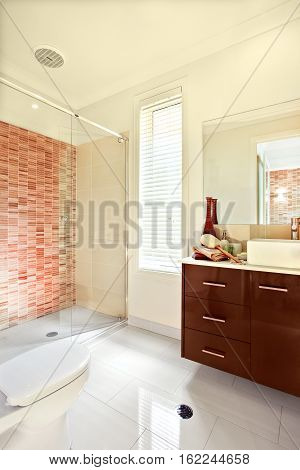 Luxury bathroom has white floor tiles exposed to the sunlight. The brown wooden cabinet has drawers and counter top with washstand in front of the mirror the wall is decorated with some color pattern
