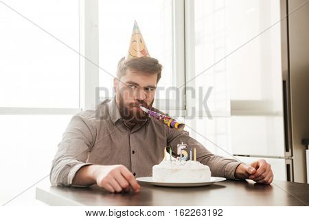 Image of angry bearded father of birtday little boy sitting in kitchen while look aside.