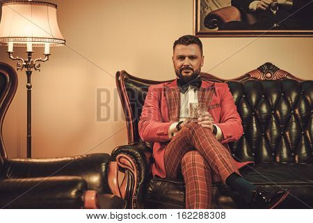 Extravagant stylish man with glass of whisky sitting on classic leather sofa in gentleman club