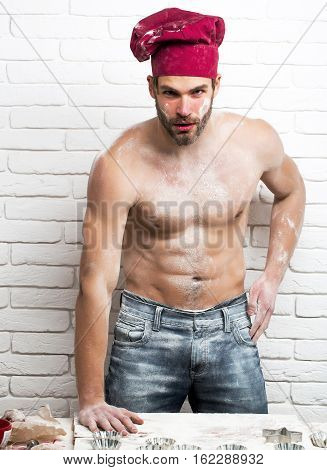 Handsome man or muscular cook baker in red chef hat poses with flour on sexy muscle torso body with six packs and abs on kitchen wall